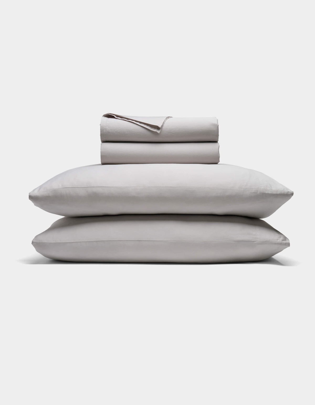 A neatly folded Soft-Wash Cotton Sheet Set by Cozy Earth in light gray is stacked atop two matching pillows, all set against a plain white background.