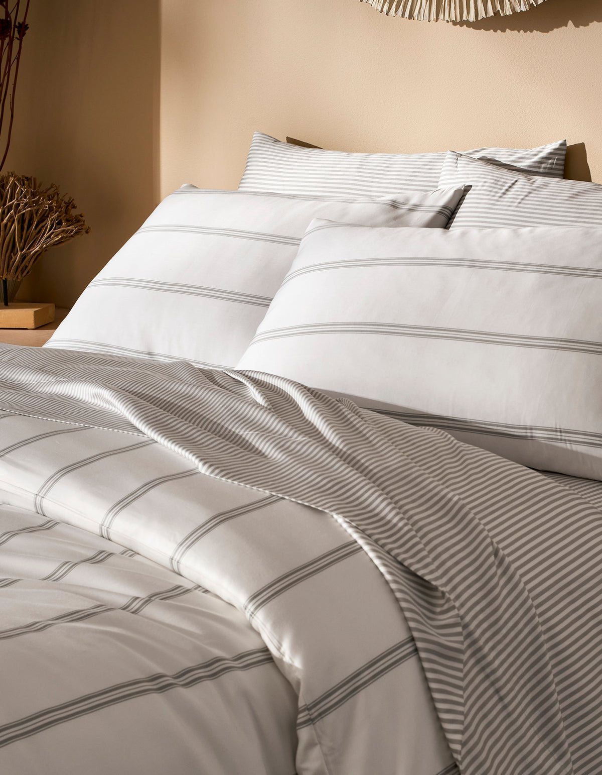 A neatly made bed features the Cozy Earth Bamboo Duvet Cover in white with light gray stripes, two large pillows, and a cozy comforter. The neutral background includes a nightstand with decorative dried plants.