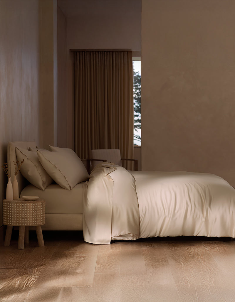 A minimal, modern bedroom features a neatly made bed with Cozy Earth's Bamboo Duvet Cover in cream, a round nightstand with a vase of dried plants, wood floors, and a window with brown curtains partially revealing greenery outside.