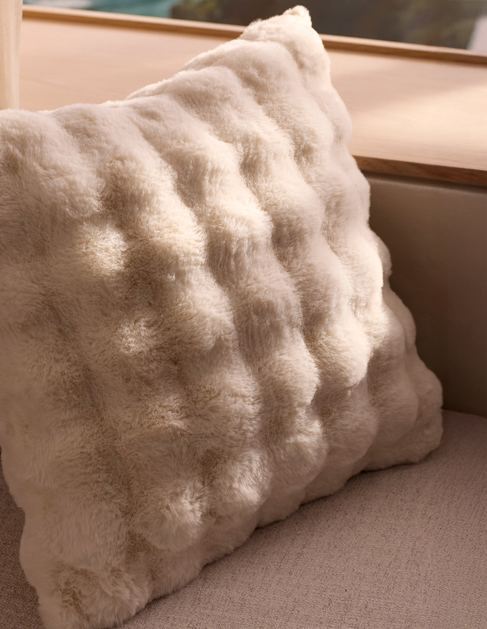 The Cozy Earth Bubble Cuddle Pillow, a soft cream faux fur with a raised texture, rests on a light beige couch by a wooden window ledge, illuminated by natural sunlight.