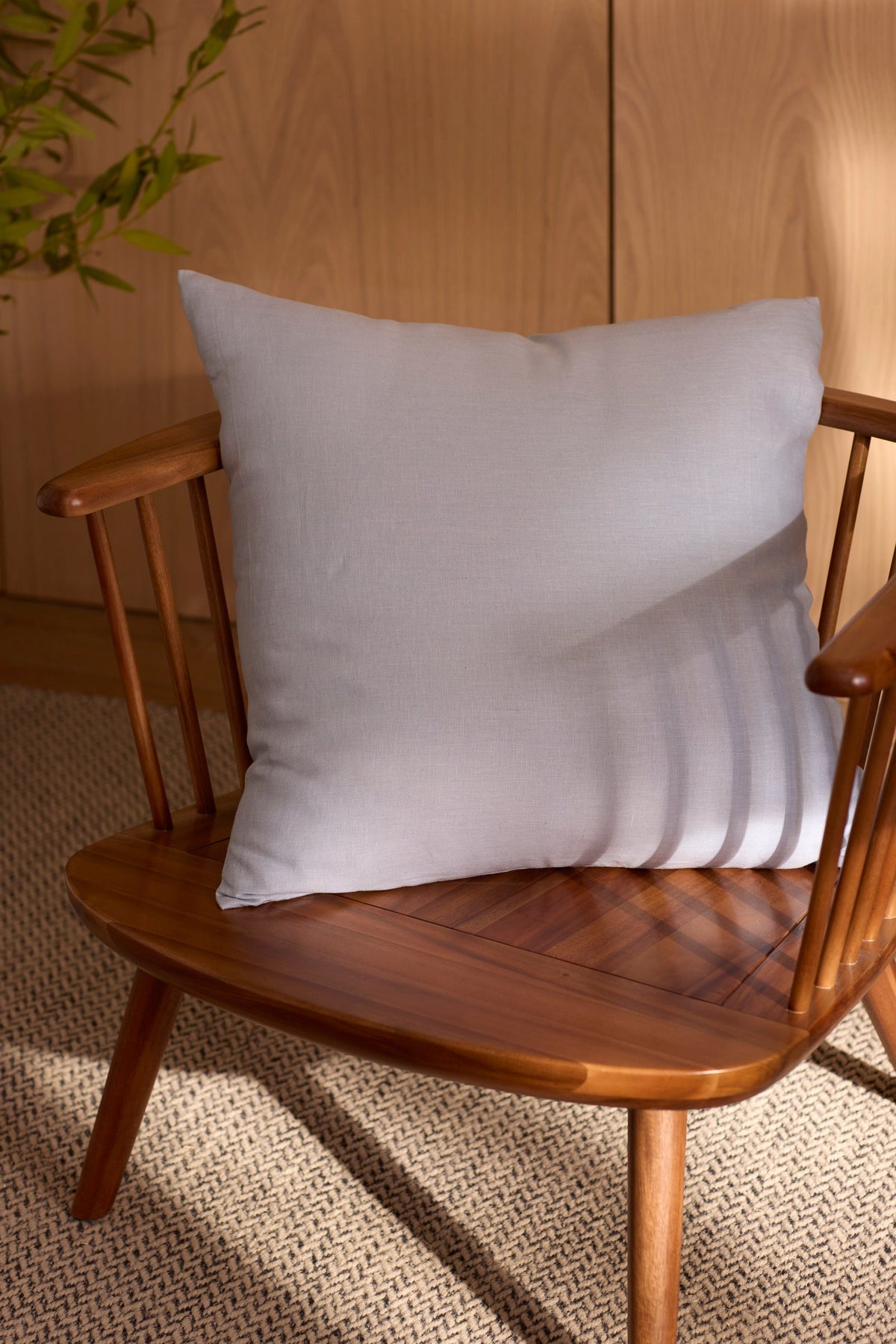 A Cozy Earth Linen Throw Pillow in light gray rests on a wooden spindle-back chair atop a textured beige rug, with sunlight and greenery in the background. 