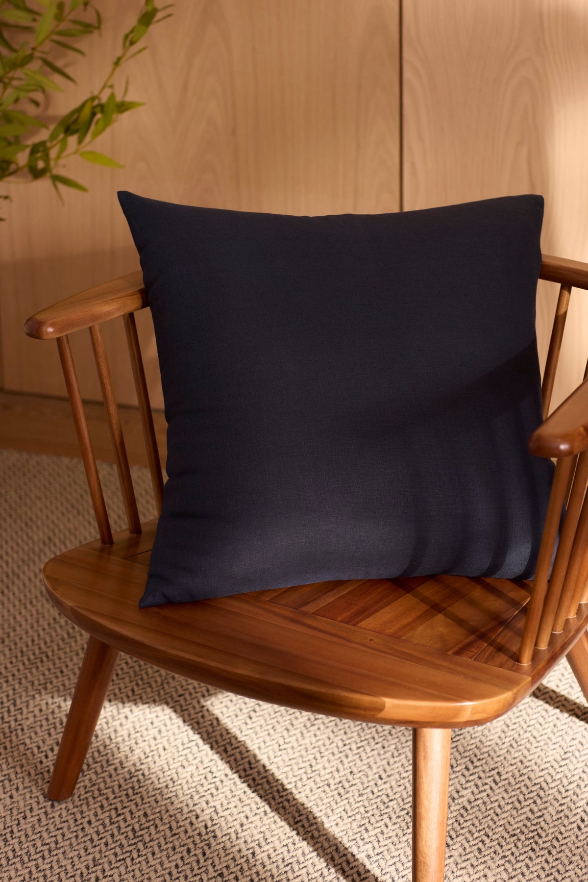 A Cozy Earth Linen Throw Pillow in dark navy blue rests on a wooden chair with spindled backrest, set atop a textured beige carpet. Sunlight casts gentle shadows while a leafy plant appears partially in the background. 