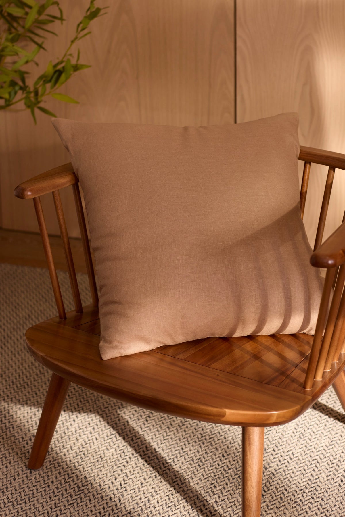 The Cozy Earth Linen Throw Pillow rests on a wooden spindle-back chair, placed on a textured rug near a wooden wall and green leafy plant, with warm sunlight casting soft shadows across the scene. 