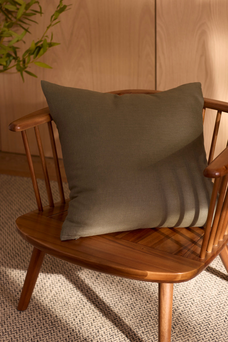 A wooden chair with spindle arms holds the Cozy Earth Linen Throw Pillow in olive green. Soft sunlight casts shadows on the pillow and chair, with a green plant and light wood paneling visible in the background.