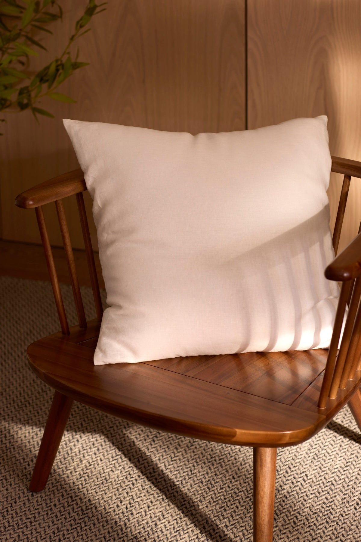 A Cozy Earth Linen Throw Pillow rests on a wooden chair with wide, angled arms, placed on a textured rug. Soft natural light casts shadows across the white pillow and wood.