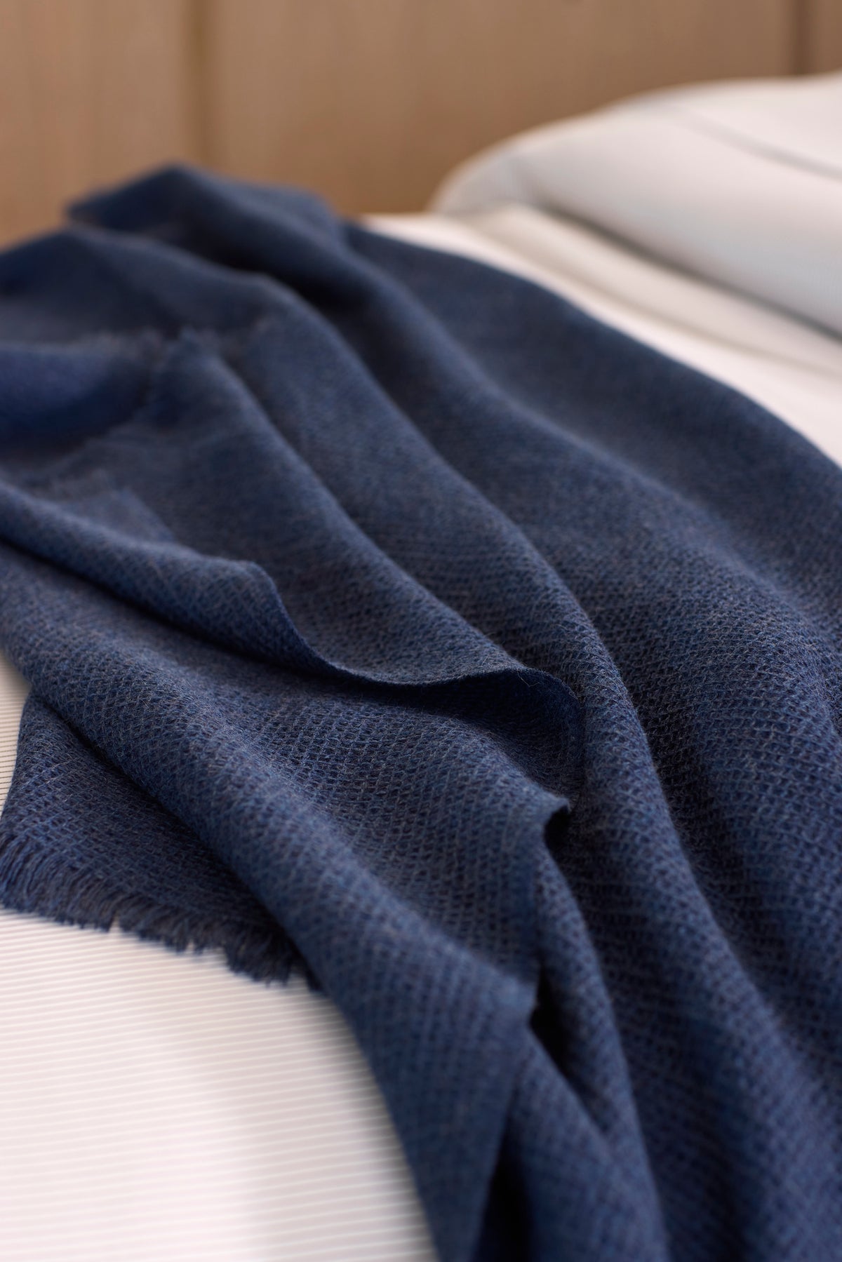 The Cozy Earth Alpaca Waffle Throw, a dark blue textured blanket with fringed edges, is draped casually over a neatly made bed with white sheets.