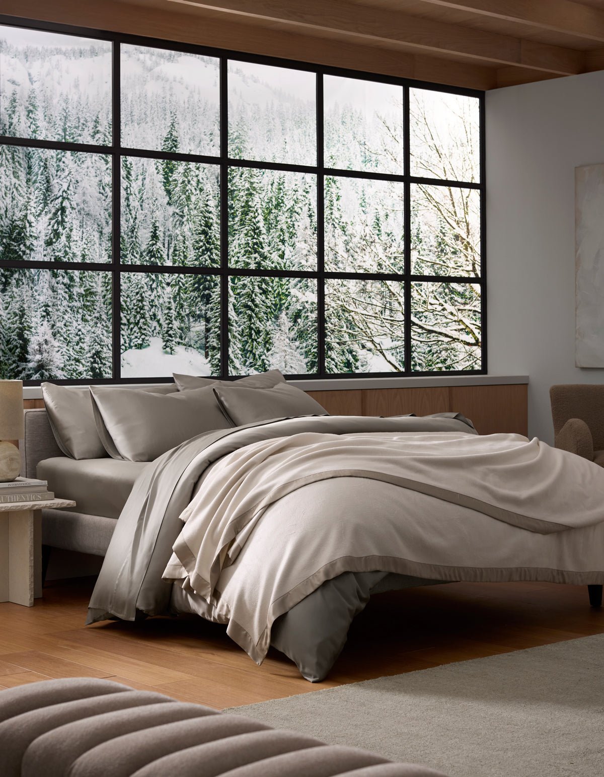 A modern bedroom features Cozy Earth's Bamboo Blanket in neutral tones on an unmade bed, large windows with snowy forest and mountain views, wooden floors, minimalist furniture, and abundant natural light for a serene, cozy feel.