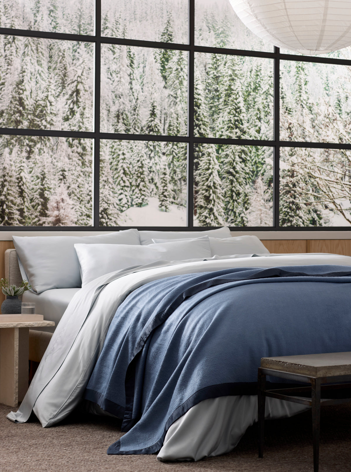 A neatly made bed, topped with a Cozy Earth Bamboo Blanket, sits before large windows revealing a snowy evergreen forest. A small table and bench are nearby, while a paper lantern hangs overhead.