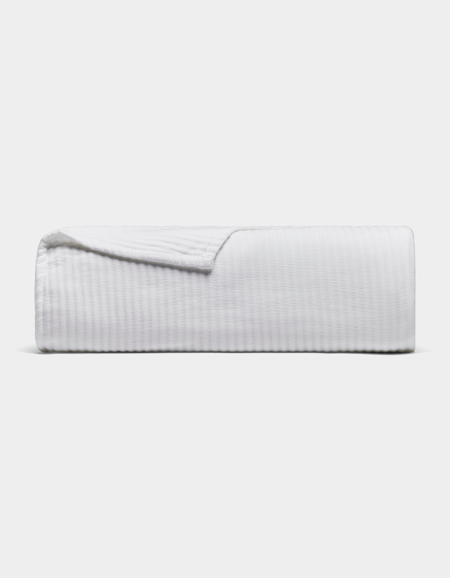 The Cozy Earth Bamboo Coverlet, a white textured blanket, is neatly folded with one corner slightly lifted, set against a plain light gray background.|Color:White