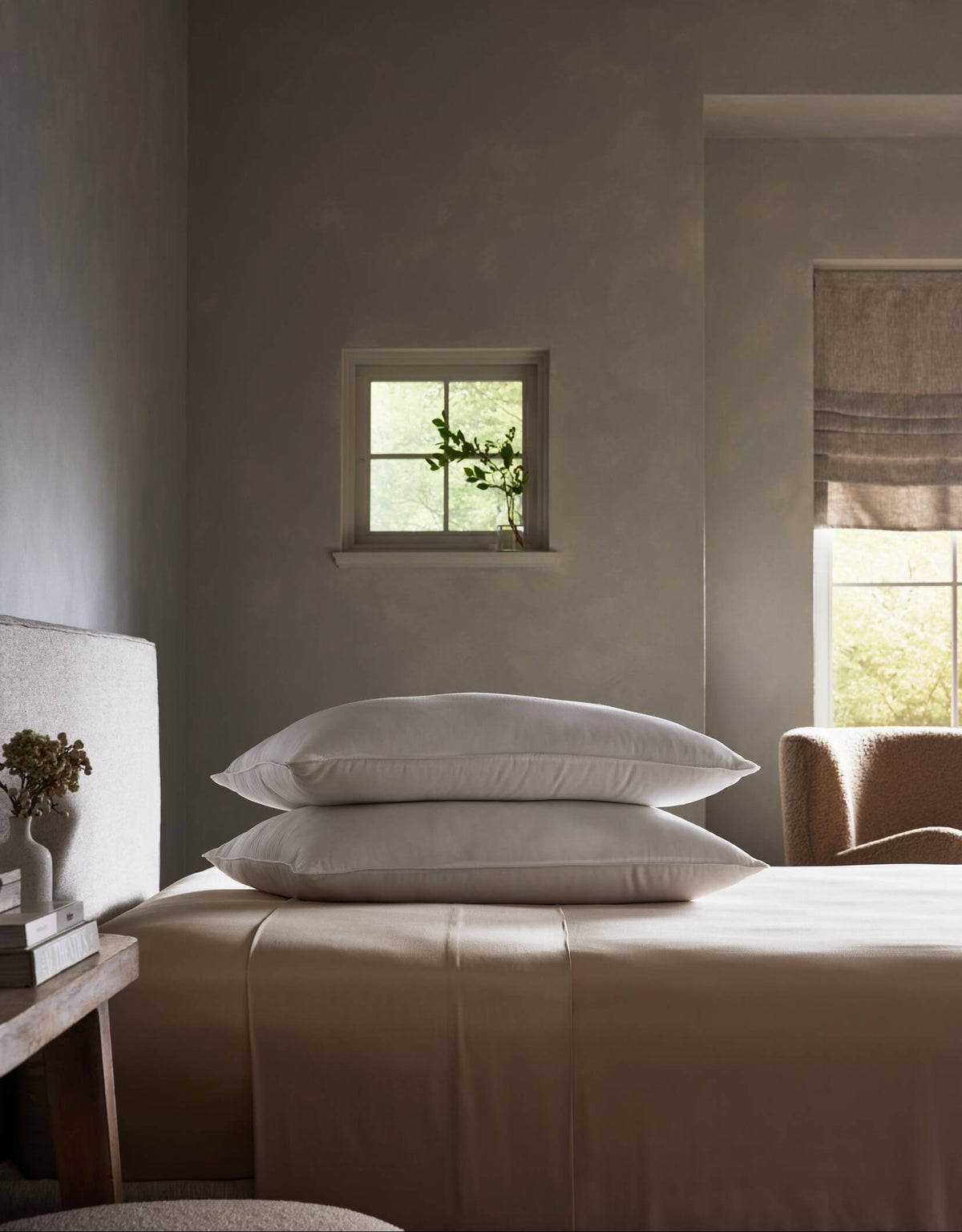 A serene, minimalist bedroom in neutral tones features a neatly made bed with the Cozy Earth Down Alternative Insert Bundle and two stacked white pillows, softly illuminated by daylight near a window with a glass vase of greenery.