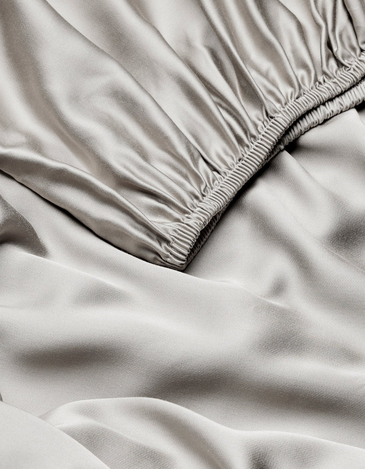 Close-up of Cozy Earth Bamboo Fitted Sheet in light gray, featuring a gathered elastic waistband and showcasing its smooth, shiny, slightly wrinkled satin texture.
