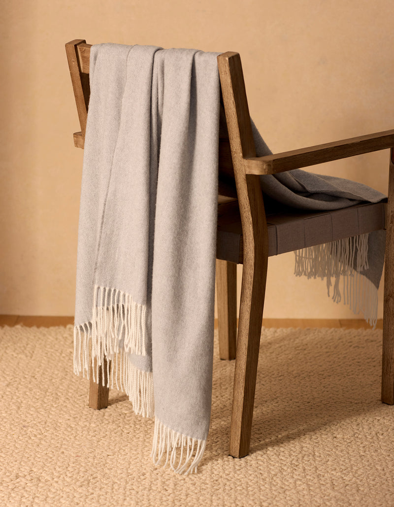 The Bamboo Herringbone Tassel Throw by Cozy Earth, in light gray with fringe, is draped over a cushioned wooden chair on a woven beige rug against a neutral wall.