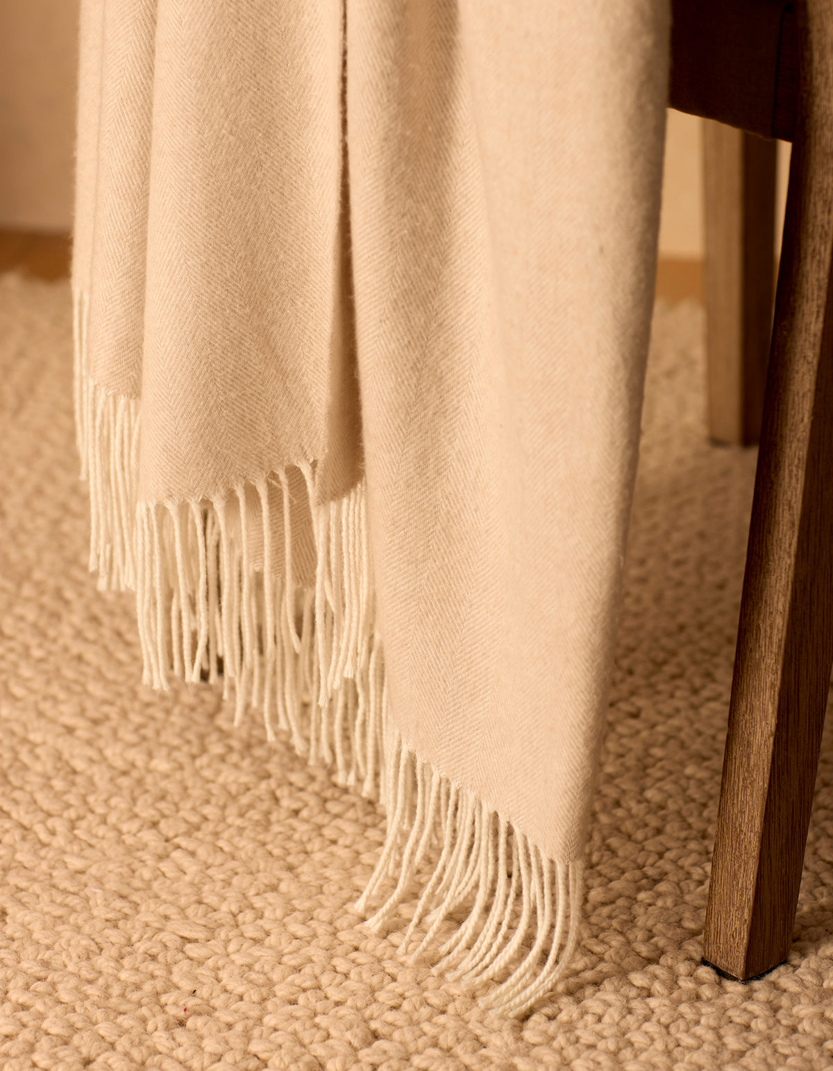 The Bamboo Herringbone Tassel Throw by Cozy Earth, in a cream color with fringe, is draped over a wooden chair on a textured beige carpet.