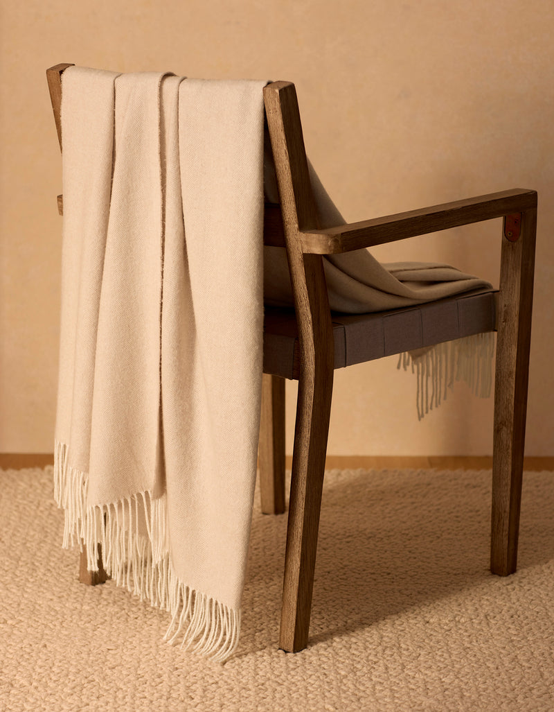 The Cozy Earth Bamboo Herringbone Tassel Throw in beige is draped over a wooden chair with a cushioned seat, set atop a textured cream carpet against a neutral wall.
