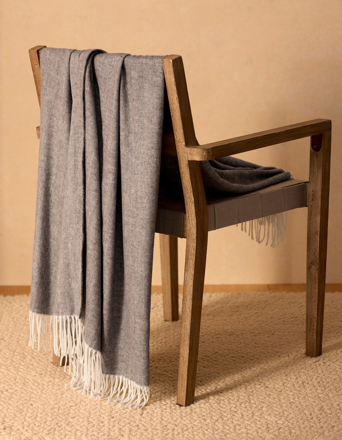 The Bamboo Herringbone Tassel Throw by Cozy Earth, in soft gray with white fringe, is draped over a wooden chair on a beige textured rug against a light tan wall. 