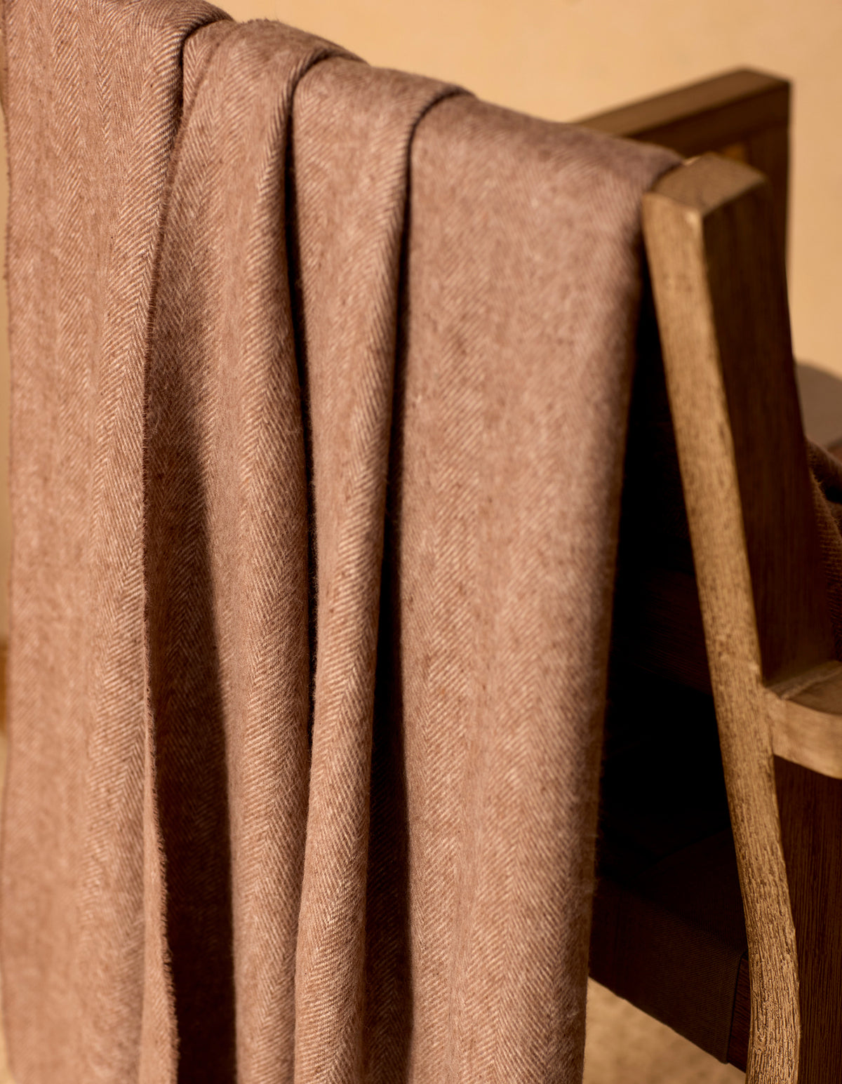 The Cozy Earth Bamboo Herringbone Tassel Throw, in light brown with a herringbone pattern, is draped neatly over a wooden chair, bathed in warm lighting.