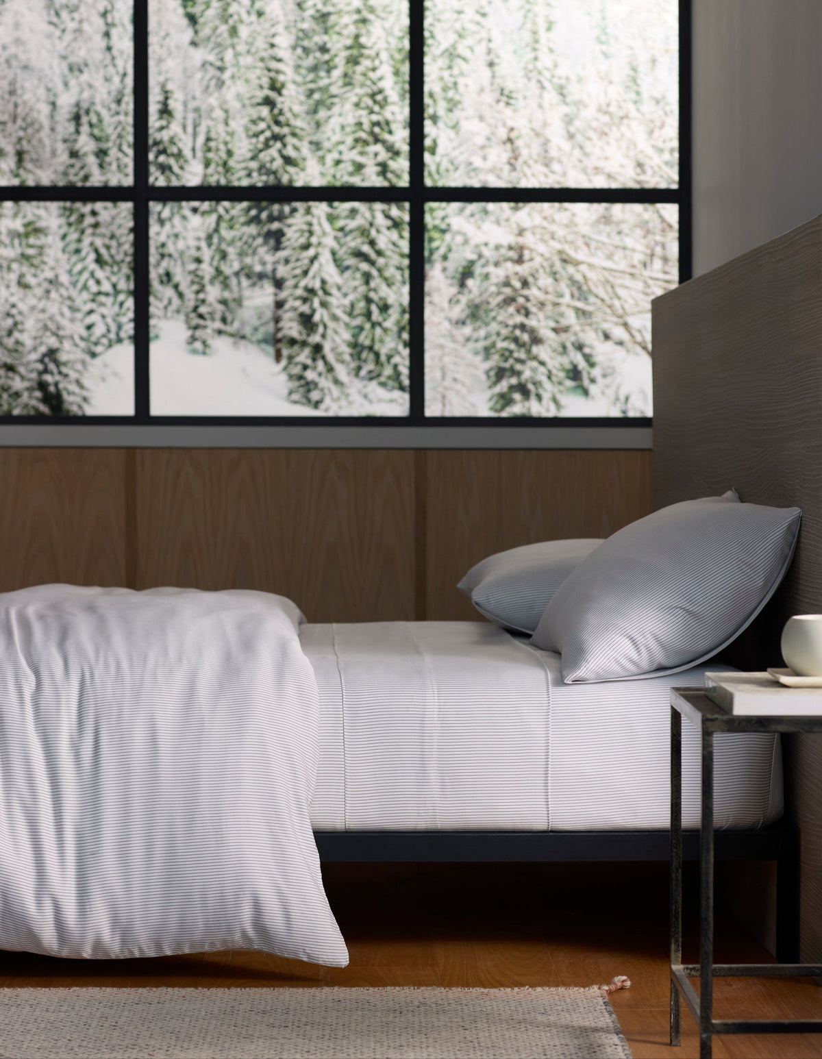 A neatly made bed with Cozy Earth’s Bamboo Sheet Set stands next to a small table with a cup, set against large windows overlooking a snowy forest scene. 