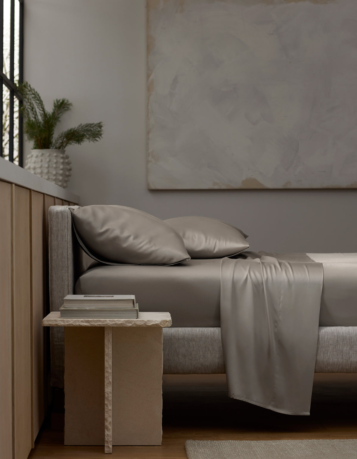 A modern, minimal bedroom features a gray upholstered bed dressed in Cozy Earth’s Bamboo Sheet Set and matching pillows. A stone nightstand holds books and a vase with greenery, while large abstract art hangs on the neutral wall.