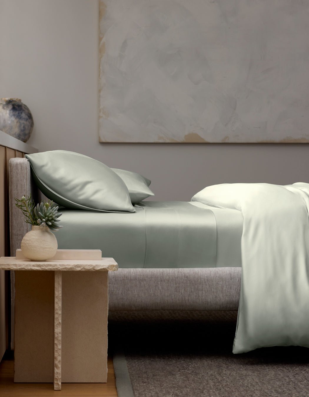 A modern bedroom featuring a neatly made bed with Cozy Earth’s Bamboo Fitted Sheet, two pillows, and a gray upholstered headboard. A small table with a potted succulent and ceramic vase sits beside the bed. Abstract art hangs on the wall.