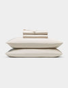 A neatly folded Cozy Earth Bamboo Sheet Set—cream-colored, including a fitted sheet, flat sheet, and two pillowcases—rests atop two matching pillows against a plain white background.|Color:Oat