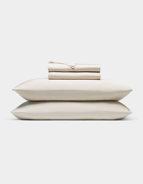 A neatly folded Cozy Earth Bamboo Sheet Set—cream-colored, including a fitted sheet, flat sheet, and two pillowcases—rests atop two matching pillows against a plain white background.|Color:Oat