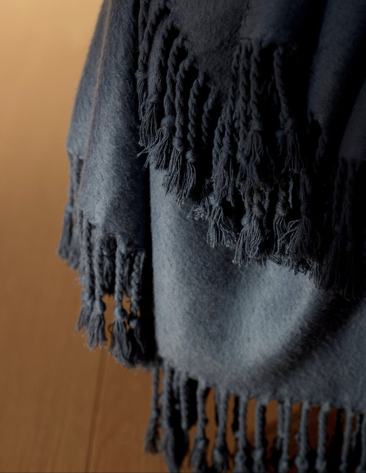 A close-up of the Cozy Earth Bamboo Tassel Throw, a dark blue blanket with fringe, draped over the edge of a wooden surface.