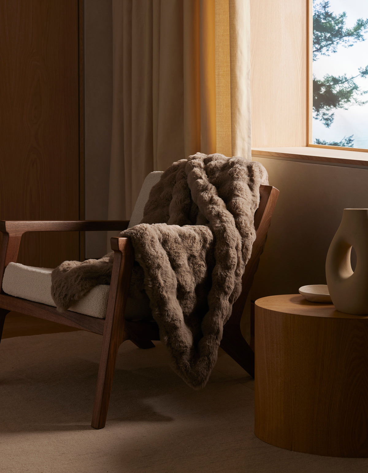 A cozy wooden armchair with a cream cushion and a Bubble Cuddle Blanket by Cozy Earth draped over it sits next to a round wooden side table with minimalist ceramic decor, beside a sunlit window with beige curtains.