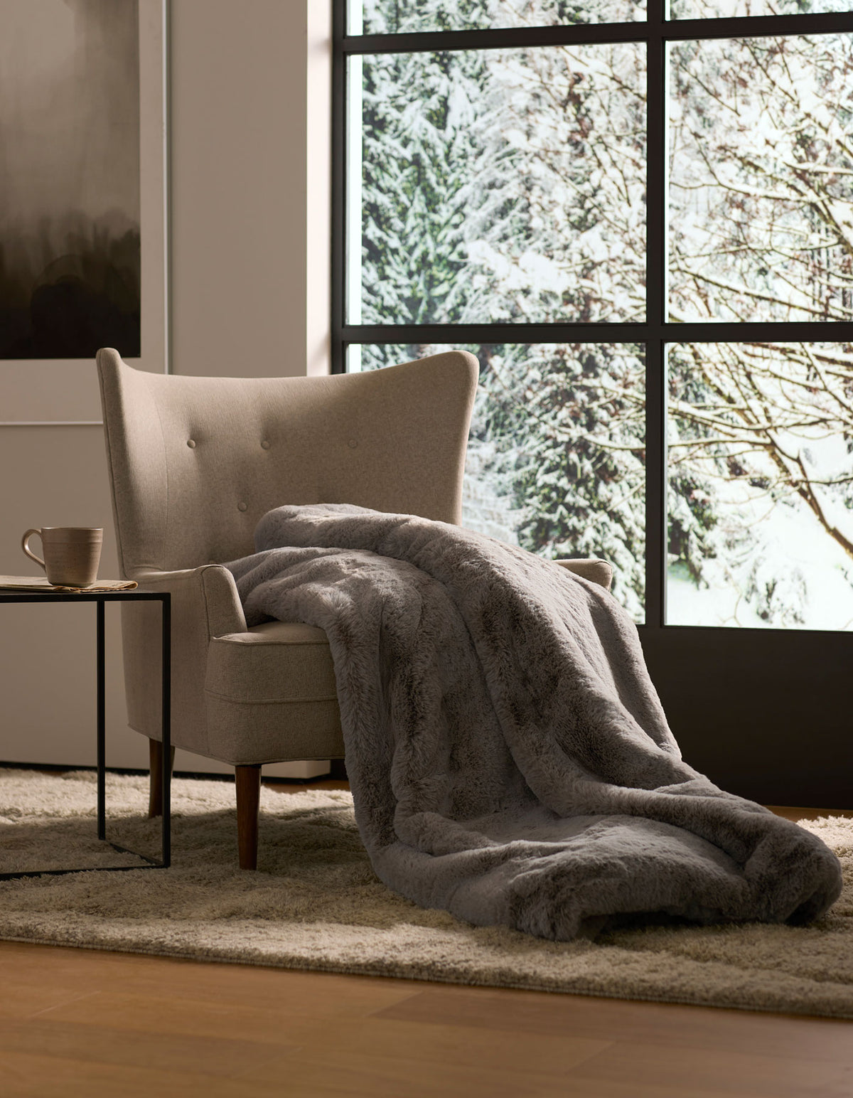 A gray armchair with a Cozy Earth Cuddle Blanket draped over it sits by a large window overlooking snowy trees, while a mug rests on the side table nearby.