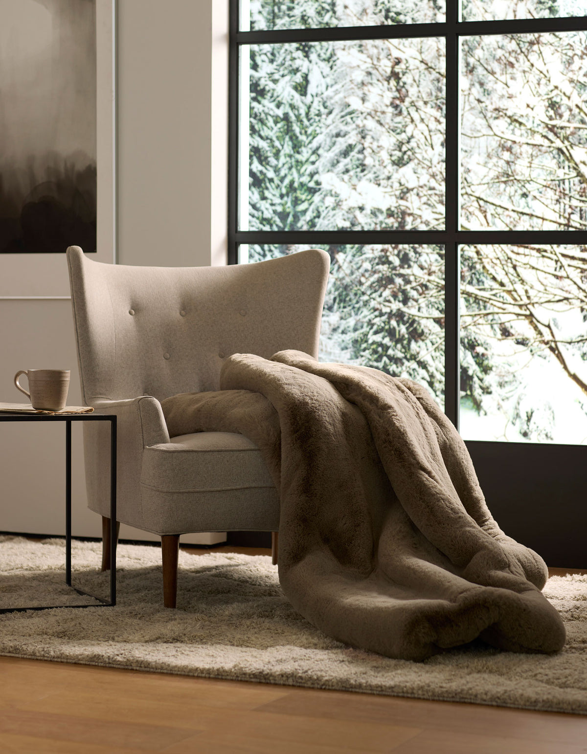 A cozy beige armchair with a Cozy Earth Cuddle Blanket draped over it sits beside a small table with a mug, in front of a large window overlooking snowy trees.