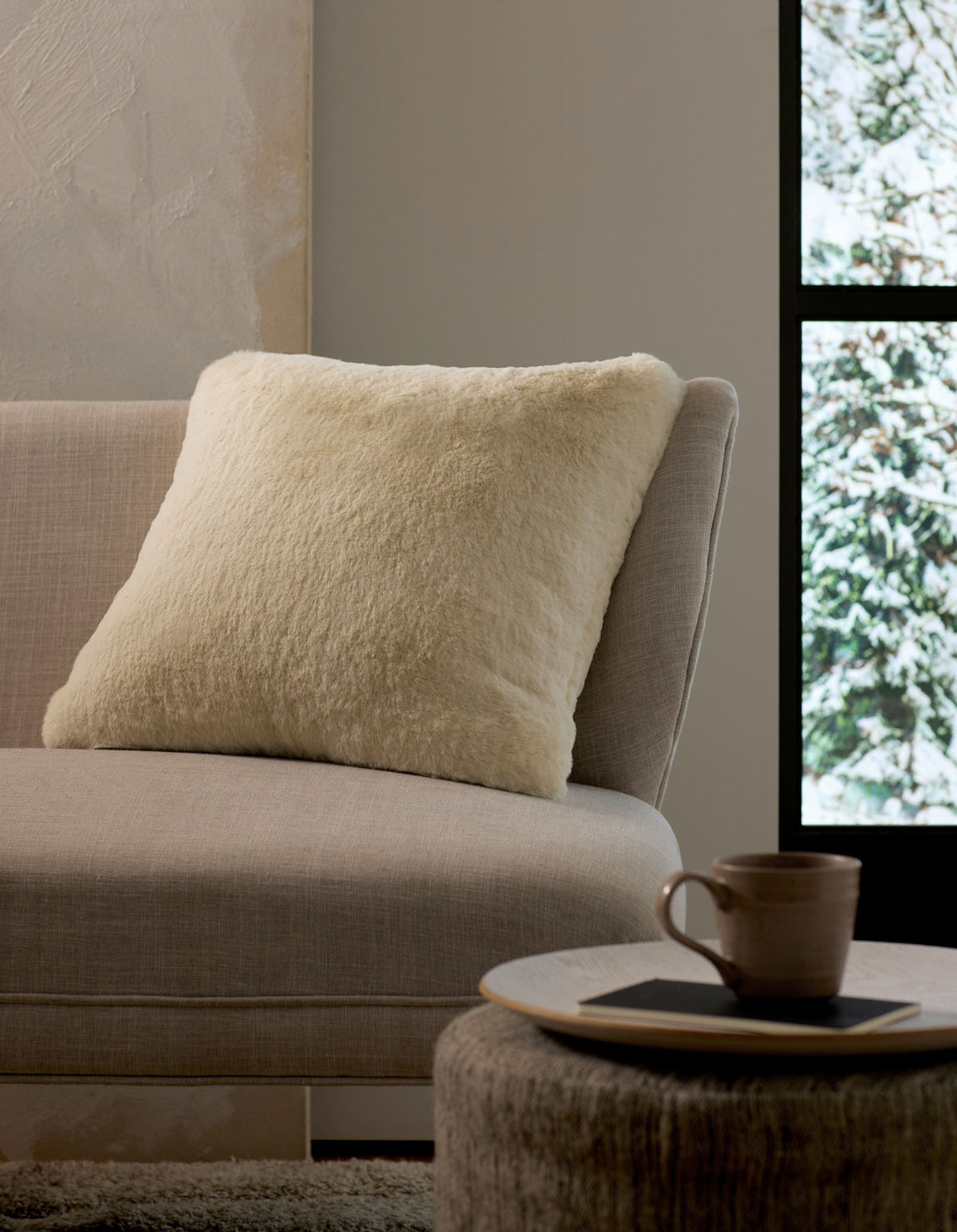 A cozy interior scene with a light beige sofa, a fluffy Cozy Earth Cuddle Pillow in cream, a round ottoman holding a cup and saucer, and a window framing a snowy outdoor view.