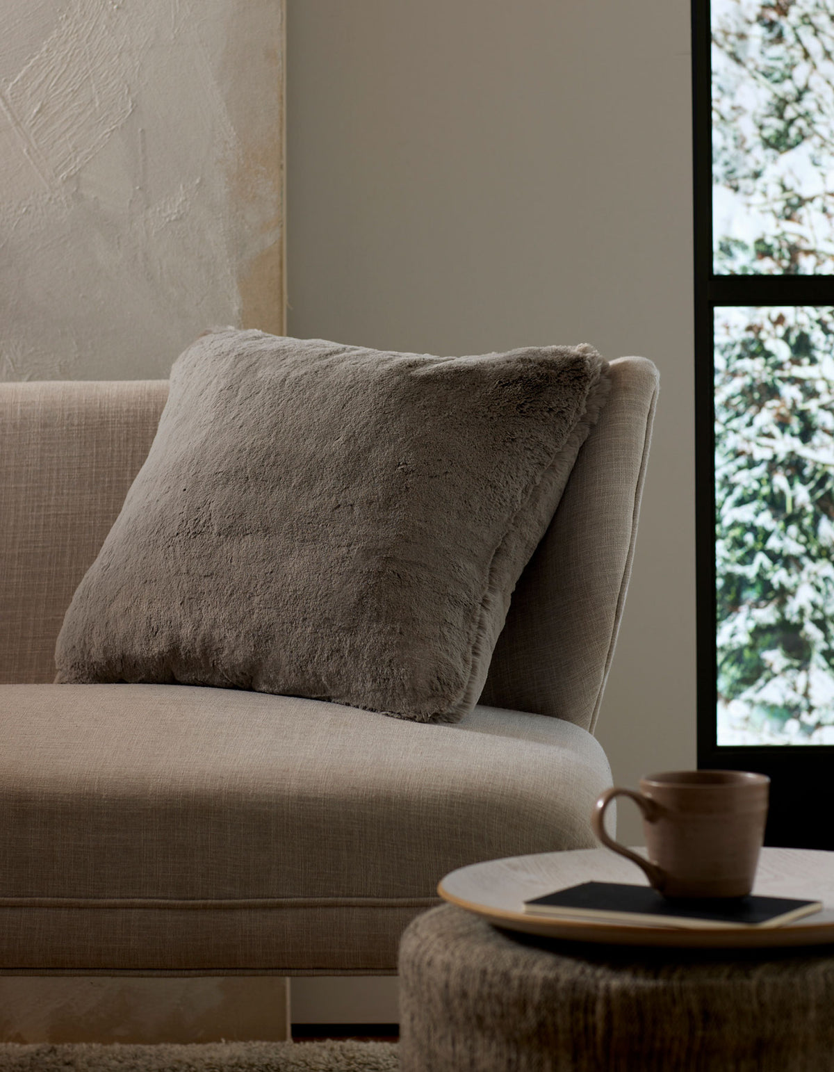 A cozy beige couch with a large, soft Cuddle Pillow by Cozy Earth rests beside a round table holding a ceramic mug and coaster. Sunlight pours through the window, revealing snowy trees outside.|Color:Dove Grey |Size:24" x 24"