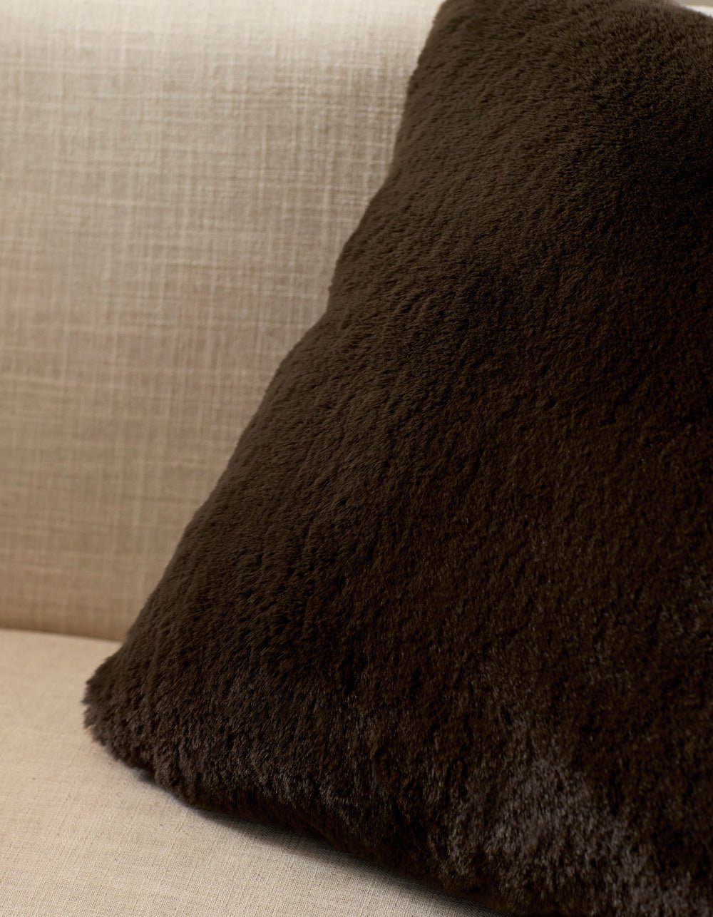 A Cozy Earth Cuddle Pillow in dark brown rests against the back of a light beige, textured fabric sofa.
