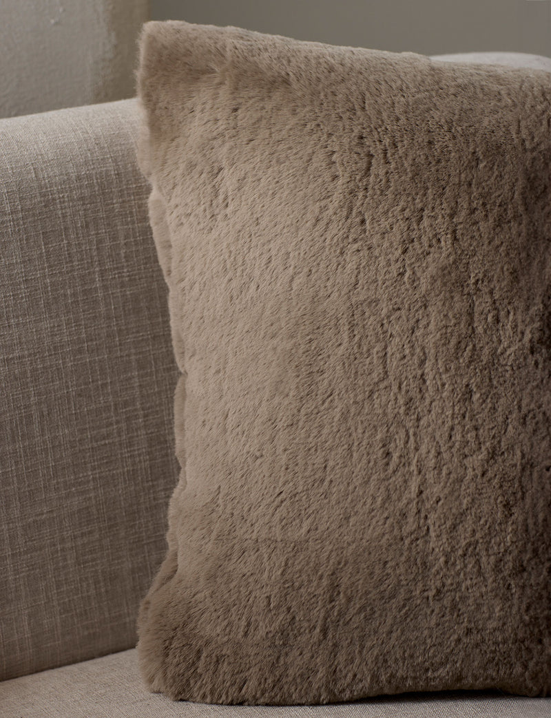 The Cozy Earth Cuddle Pillow, a square beige fuzzy throw, sits on a light fabric sofa, adding cozy softness to your neutral-toned space.