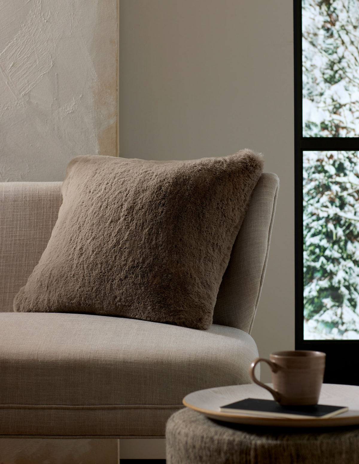 A cozy, neutral-toned living space features the Cozy Earth Cuddle Pillow in taupe on a beige sofa next to a round side table with a ceramic mug and book, framed by a window view of snowy trees.