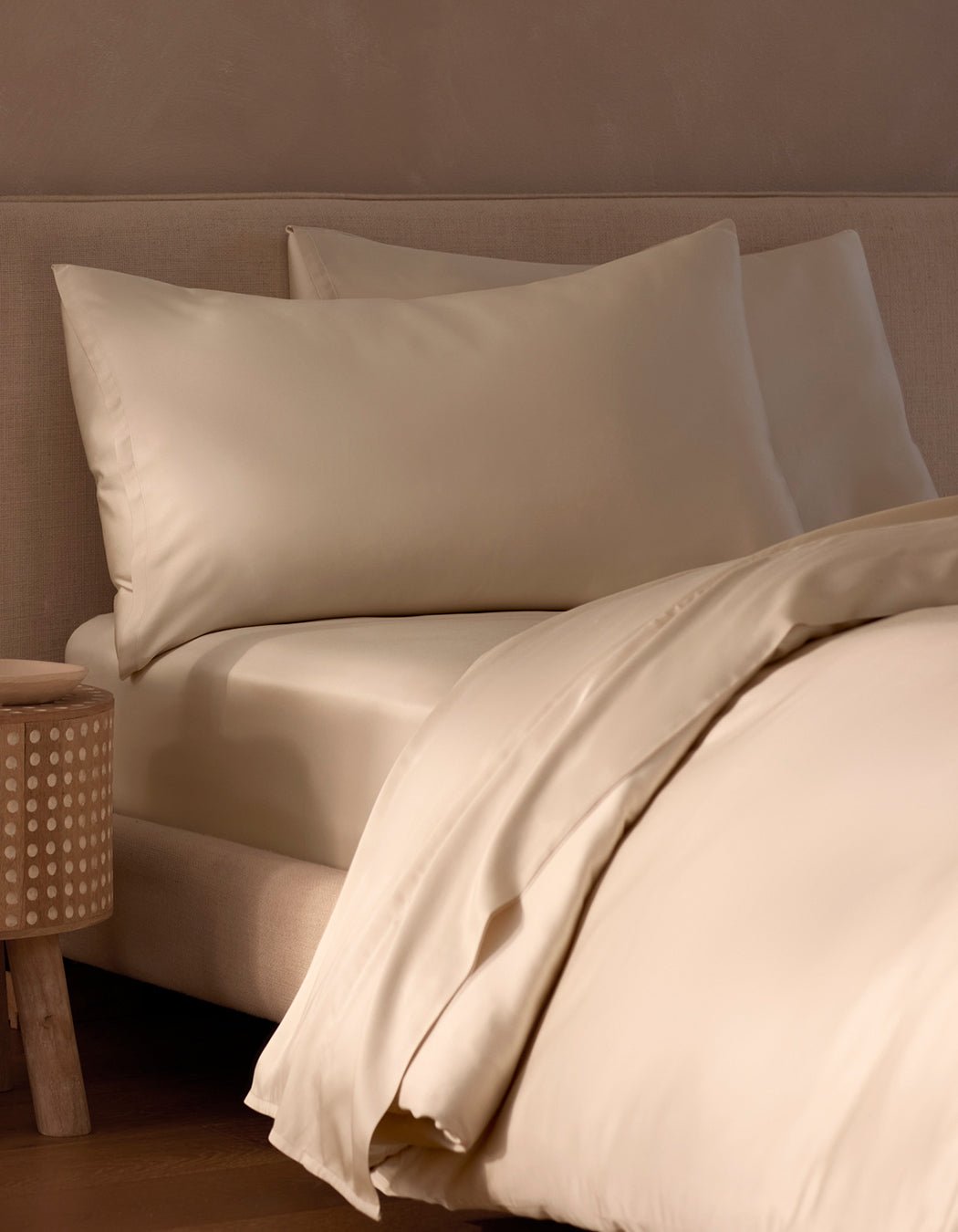 A cozy, minimalistic bedroom features a neatly made bed with cream-colored Cozy Earth Bamboo Pillowcases, sheets, and duvet, placed beside a wooden nightstand with a dotted pattern.