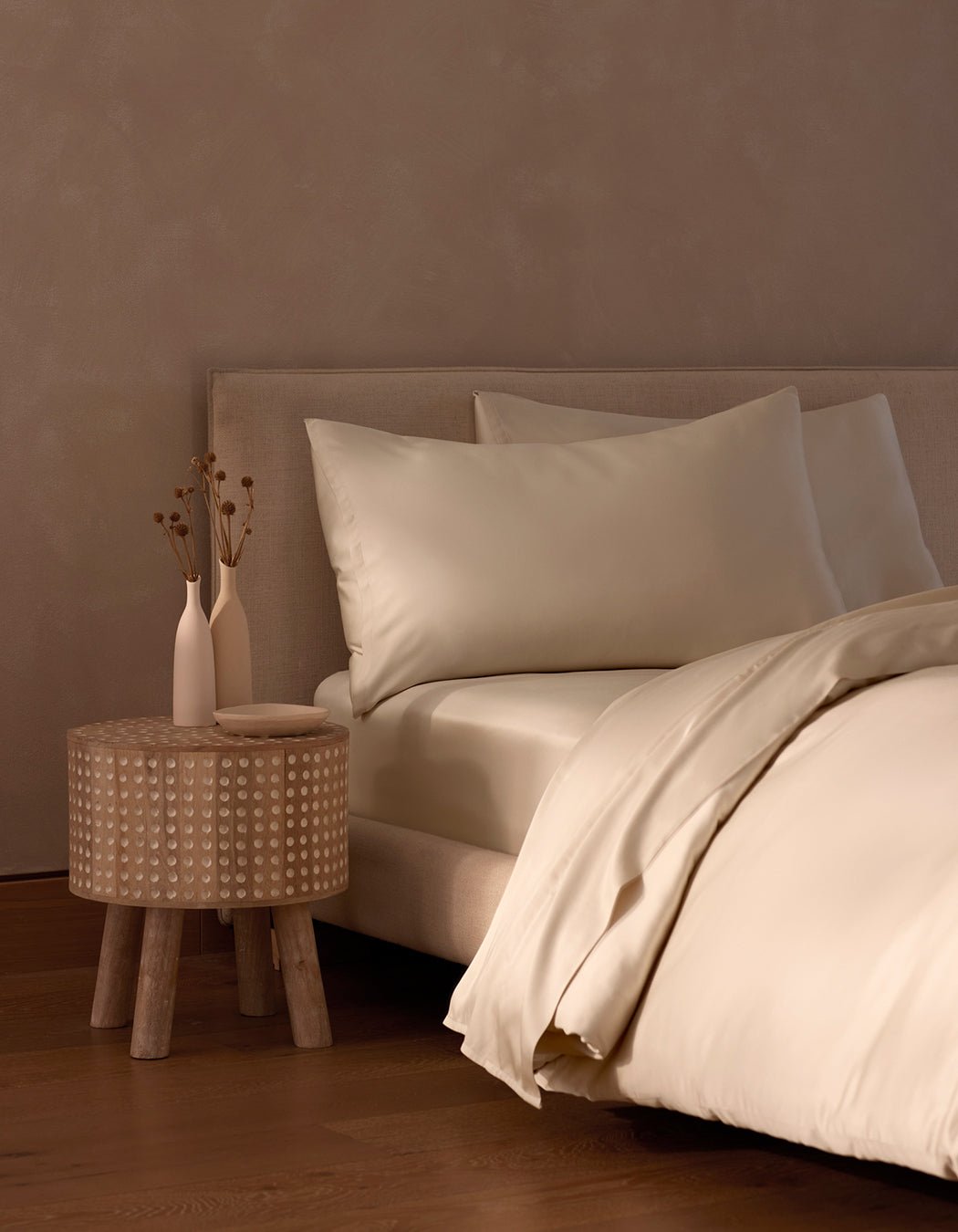 A neatly made bed with Cozy Earth’s Bamboo Bedding Core Bundle sits beside a small wooden nightstand holding two white vases with dried flowers. Cream-colored sheets and pillows complete the room’s minimalist, neutral-toned decor.|Color:Creme