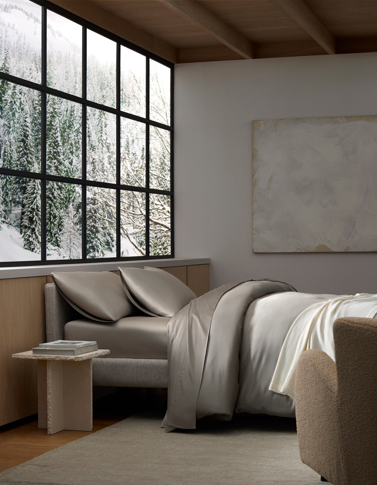 A cozy bedroom featuring Cozy Earth's Bamboo Bedding Deluxe Bundle, a gray bed and chair by the window with snowy forest views. Neutral tones, a soft rug, and a minimalistic painting create a serene, modern vibe.