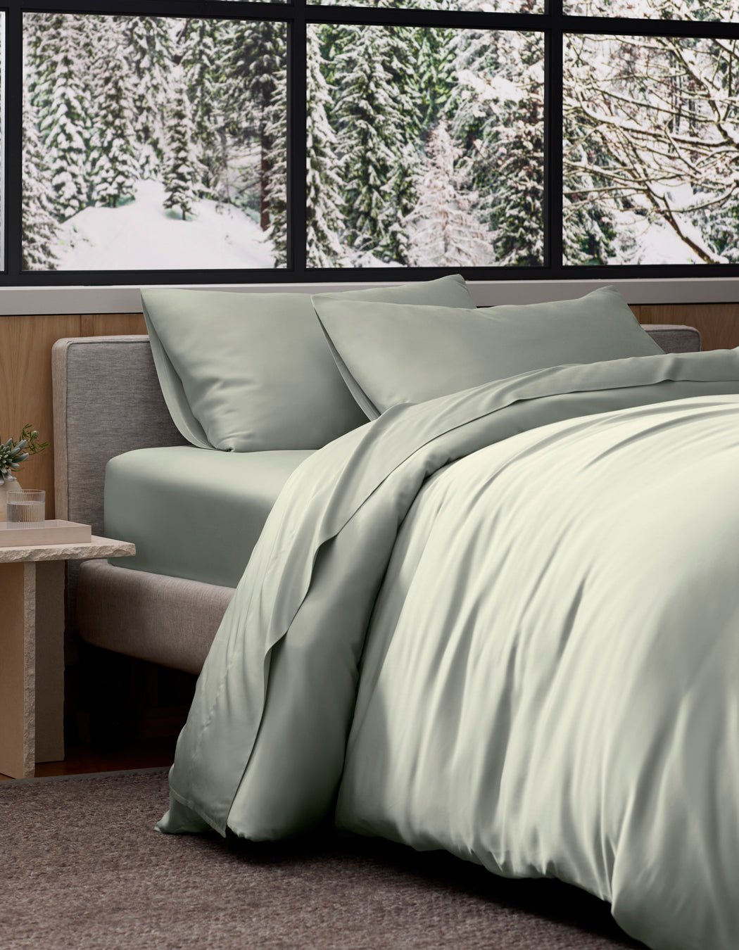 A neatly made bed features Cozy Earth's Bamboo Bedding Deluxe Bundle in light sage green, positioned by a window overlooking a snowy evergreen forest, creating a modern and cozy ambiance.