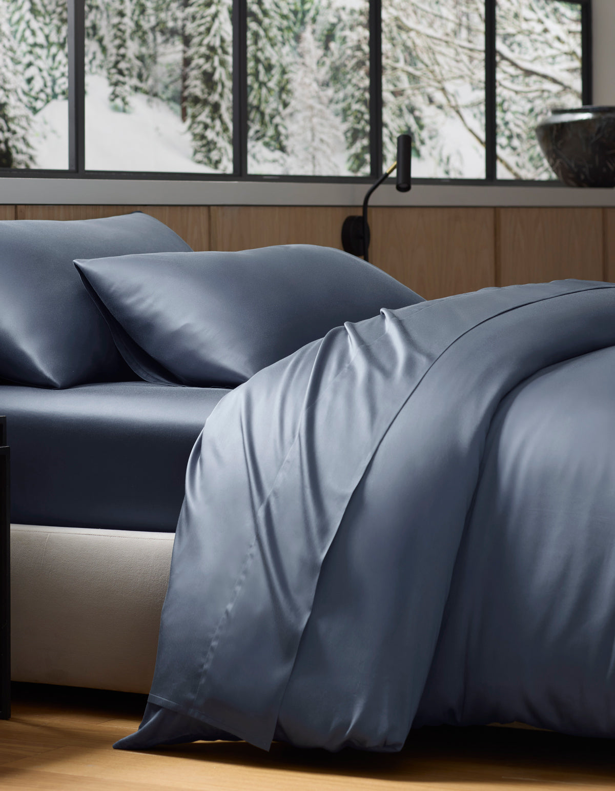 A neatly made bed with smooth, dark blue Cozy Earth Bamboo Duvet Cover and pillows sits by a large window, overlooking snow-covered trees and a wintry landscape.