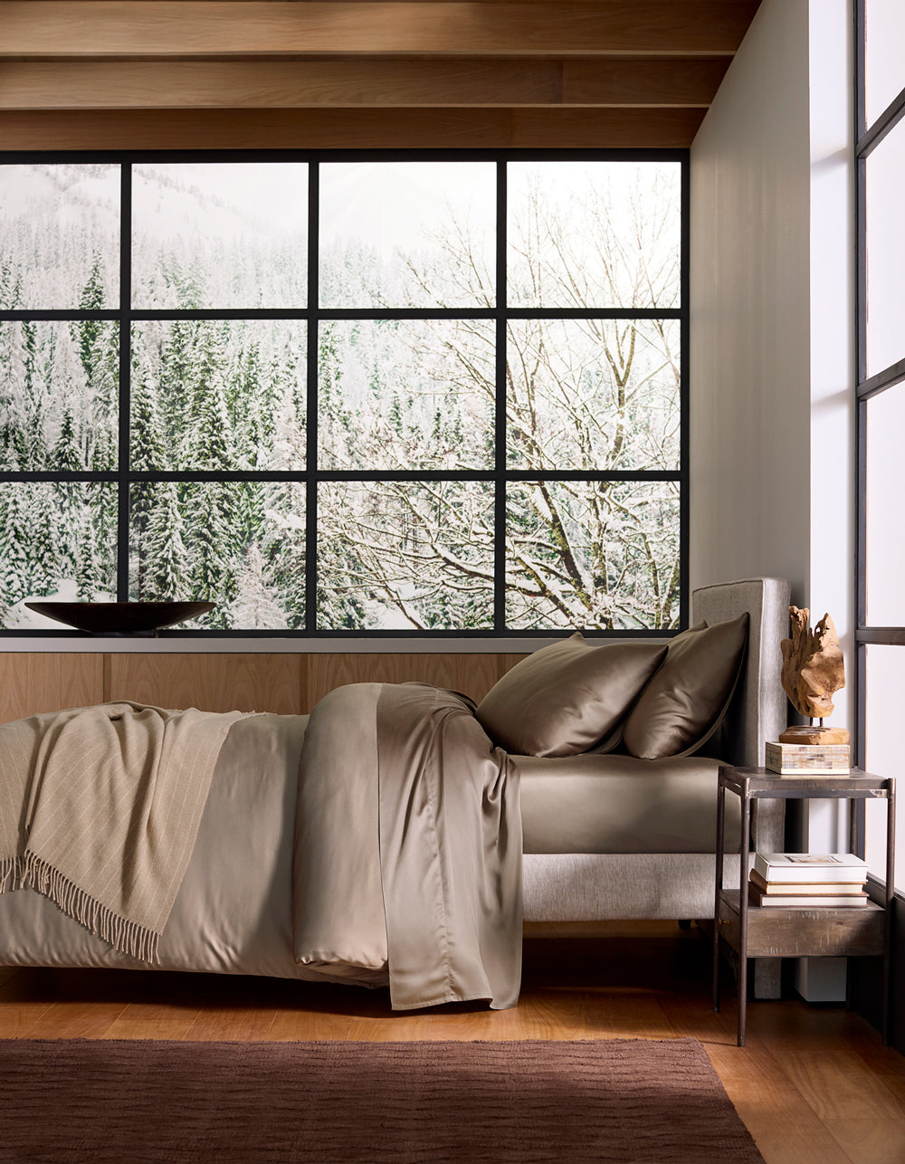 A cozy bedroom featuring a Cozy Earth Bamboo Duvet Cover with neutral tones and a throw blanket, set beside a modern nightstand and a large window revealing a snowy forest and mountain landscape outside.