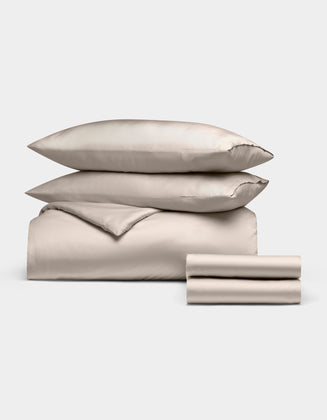 The Cozy Earth Bamboo Bedding Deluxe Bundle features two stacked pillows, a comforter, and folded sheets in cream, all neatly arranged against a plain white background.|Color:Driftwood