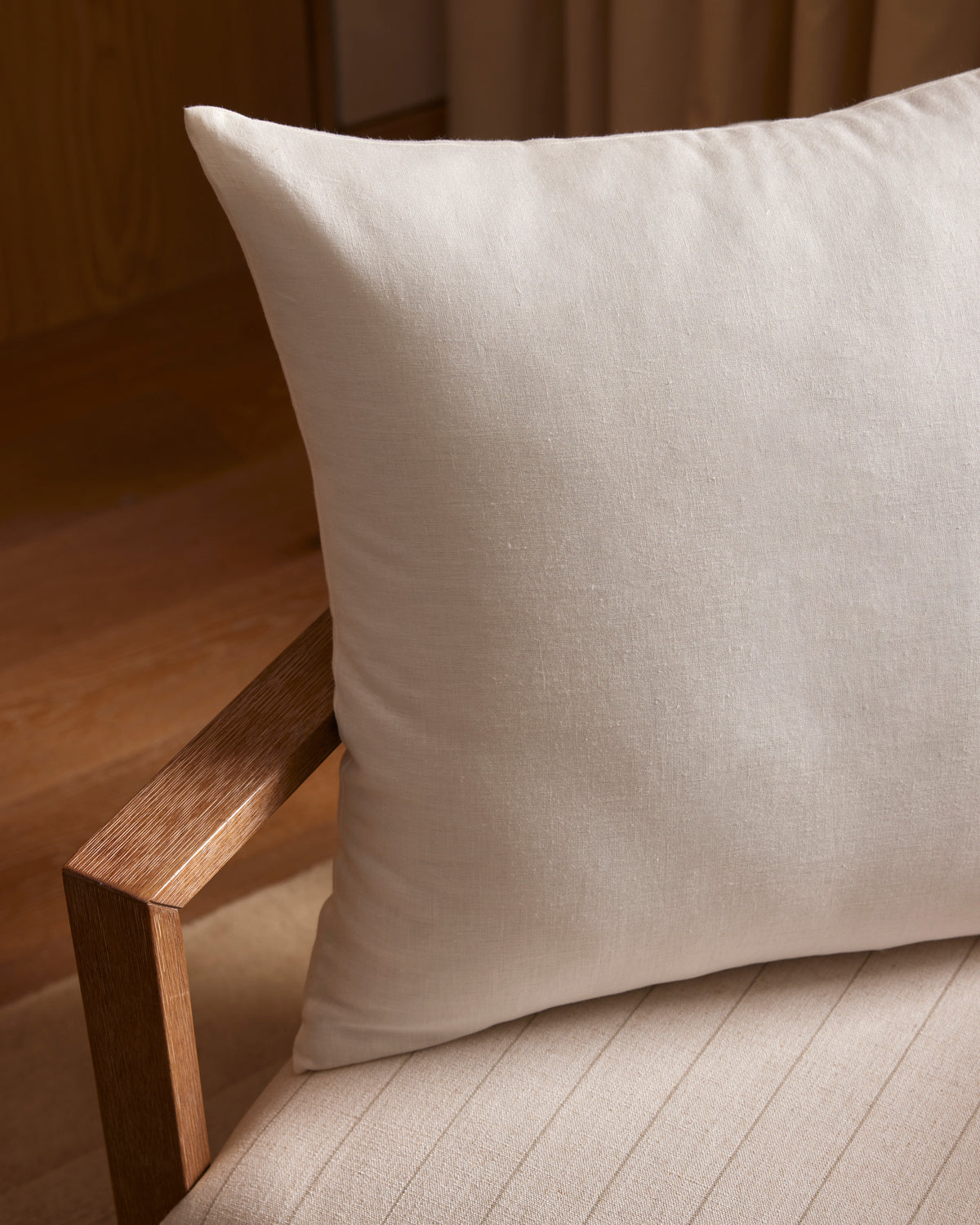 A close-up of the Cozy Earth Linen Throw Pillow on a wooden armchair with a matching striped beige cushion, set in a softly lit room with wood and fabric accents.