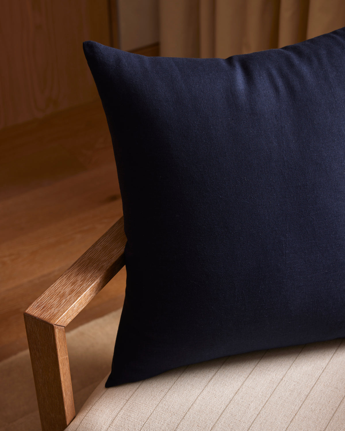 A Cozy Earth Linen Throw Pillow in dark navy rests on a light, cushioned wooden chair with clean lines. Warm wooden flooring and beige curtains create an inviting backdrop.