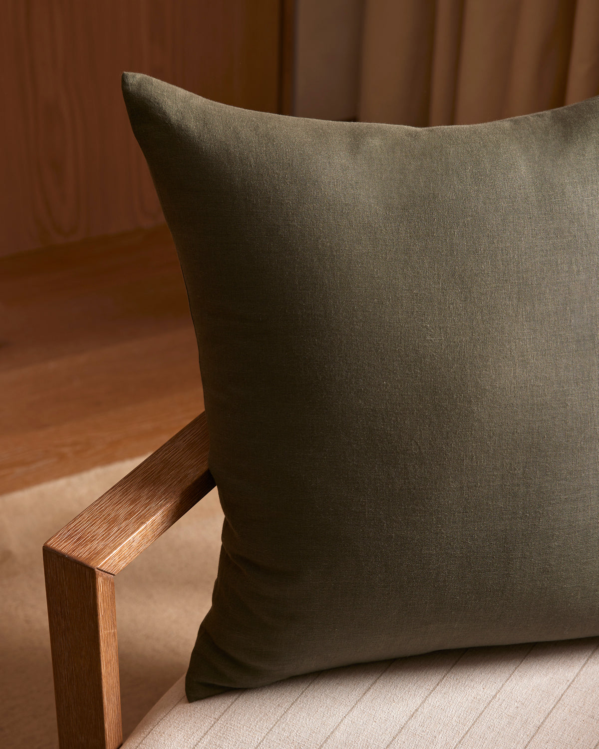 A Cozy Earth Linen Throw Pillow in dark green sits on a light beige cushioned chair with a wooden armrest, surrounded by warm lighting and wood and fabric accents in the background.