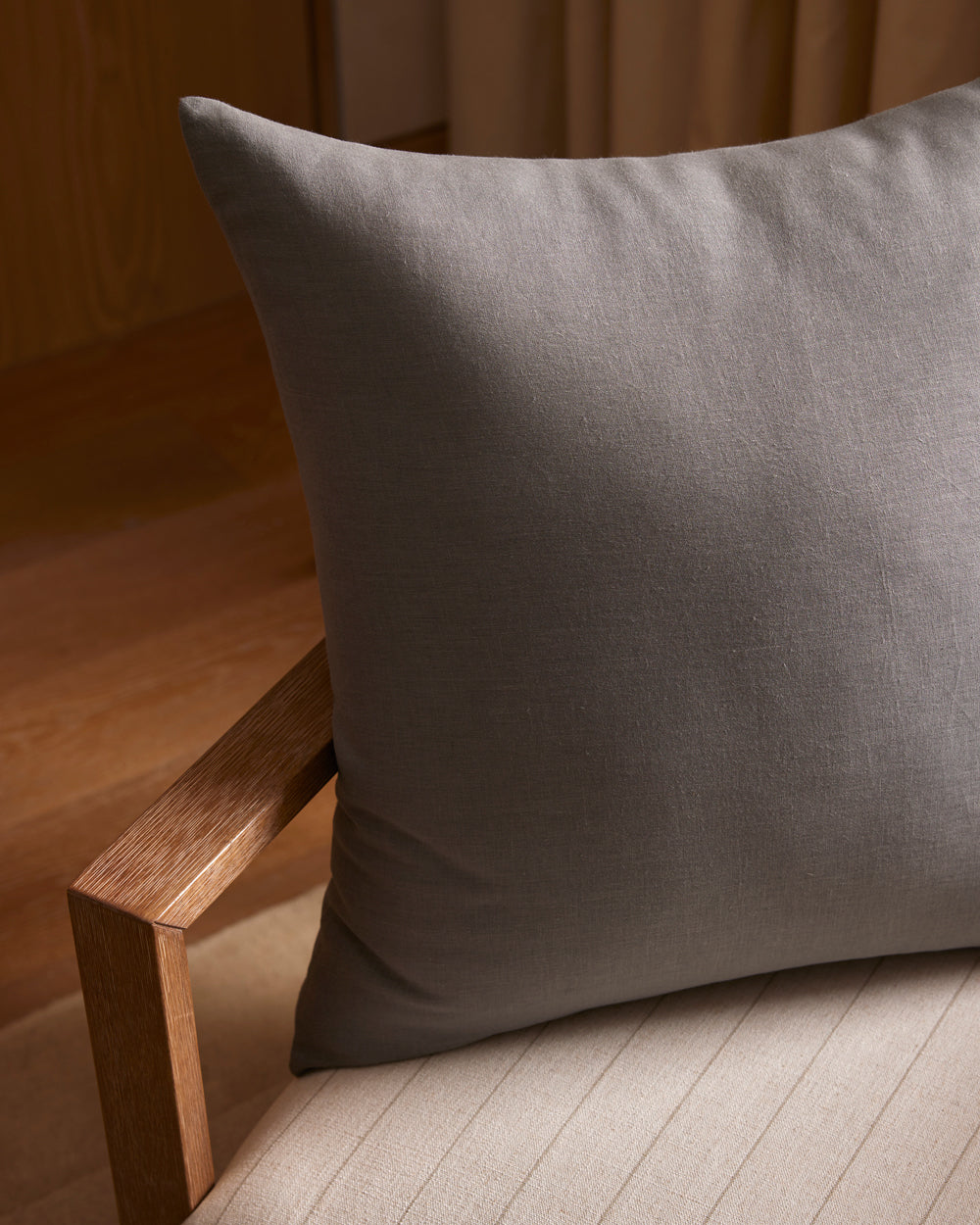 A Cozy Earth Linen Throw Pillow in gray rests on a light beige cushioned chair with wooden armrests, set in a warmly lit room featuring wood floors and neutral curtains.