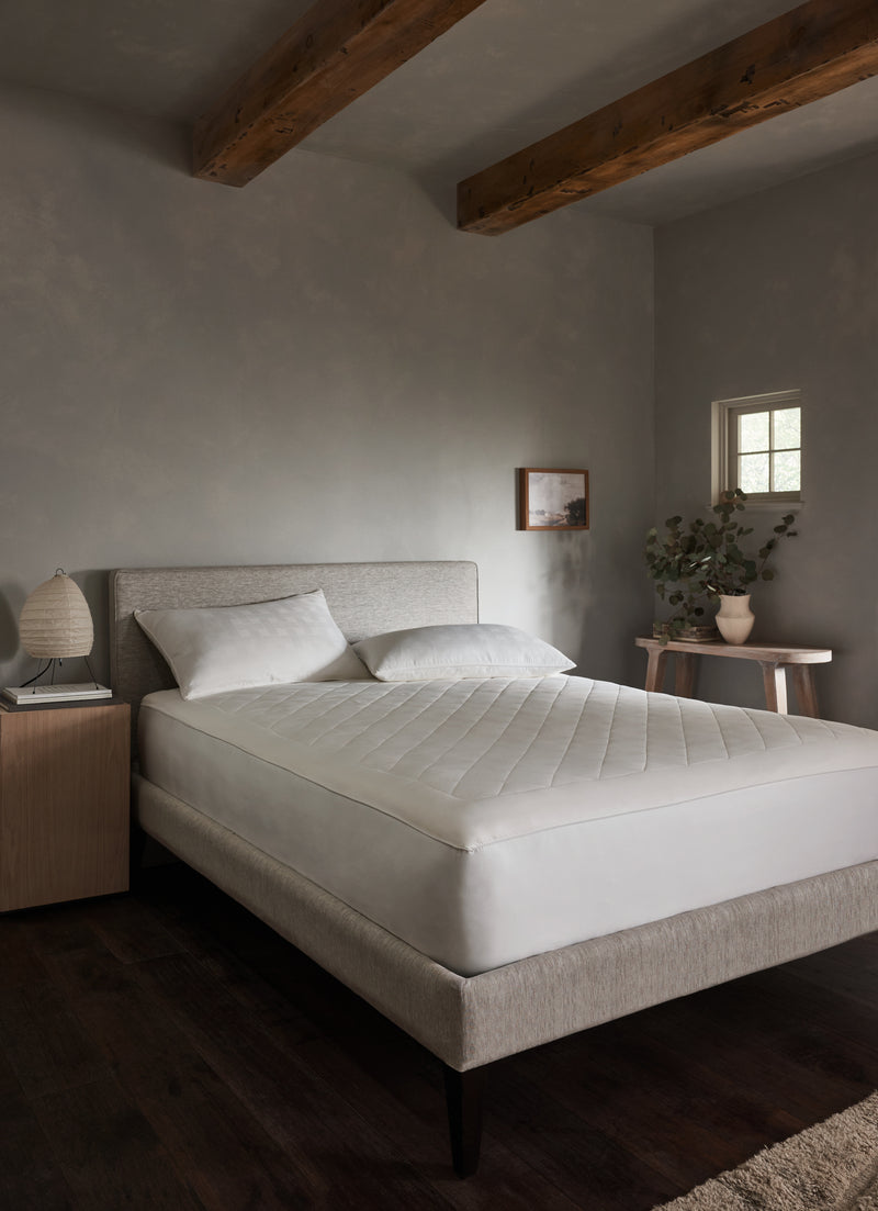 A minimalist bedroom features a light gray upholstered bed with white bedding topped by the Cozy Earth Bamboo Mattress Pad, wooden ceiling beams, dark wood flooring, a small nightstand with lamp, a table with greenery, and a small window on the wall.