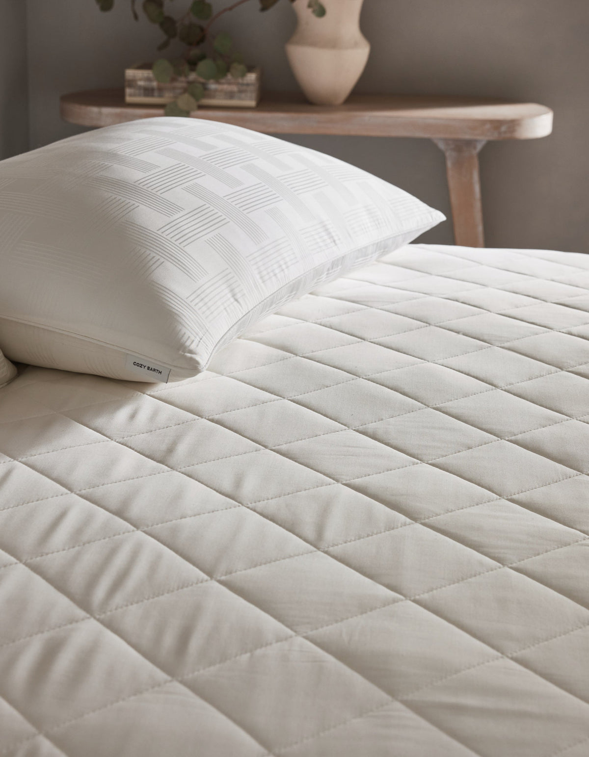 A neatly made bed features the Cozy Earth Bamboo Mattress Pad and a white pillow with a geometric pattern. In the background, a wooden table holds a ceramic vase with greenery.