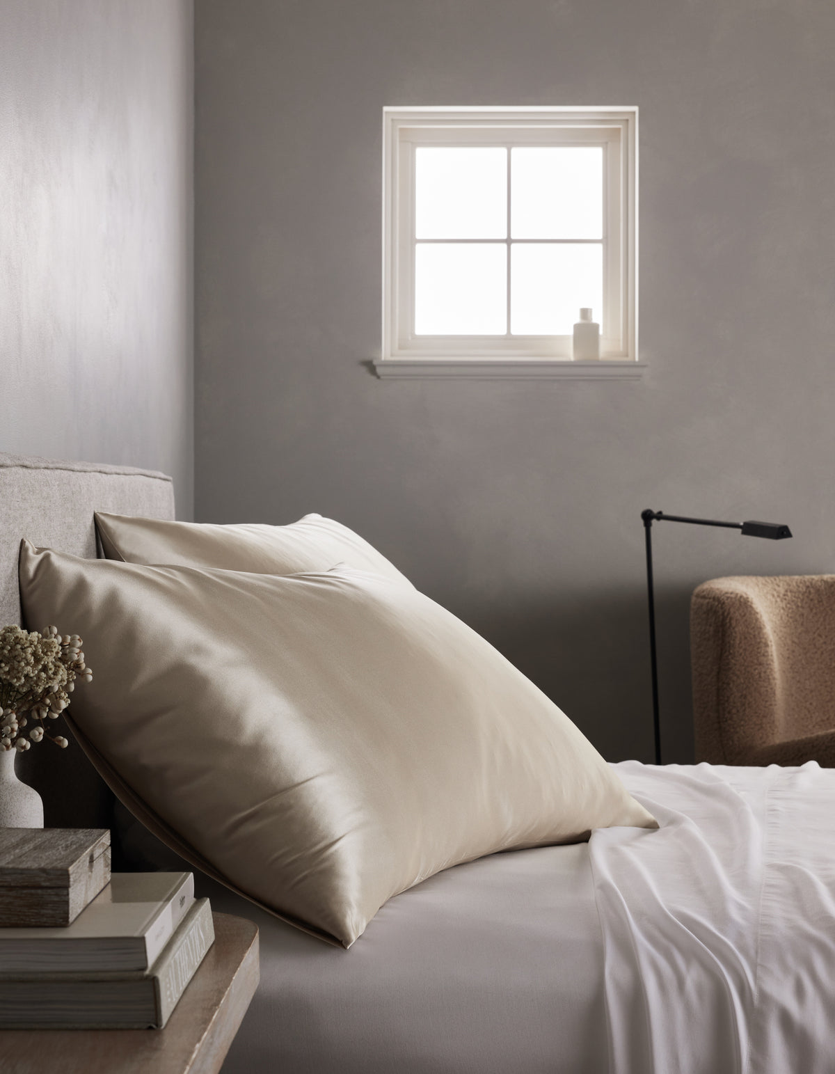 A minimalist bedroom features a neatly made bed with Cozy Earth's Silk Pillowcases, large cream-colored pillows, a nightstand holding books and dried flowers, soft light from a small window, and a beige chair in the background.