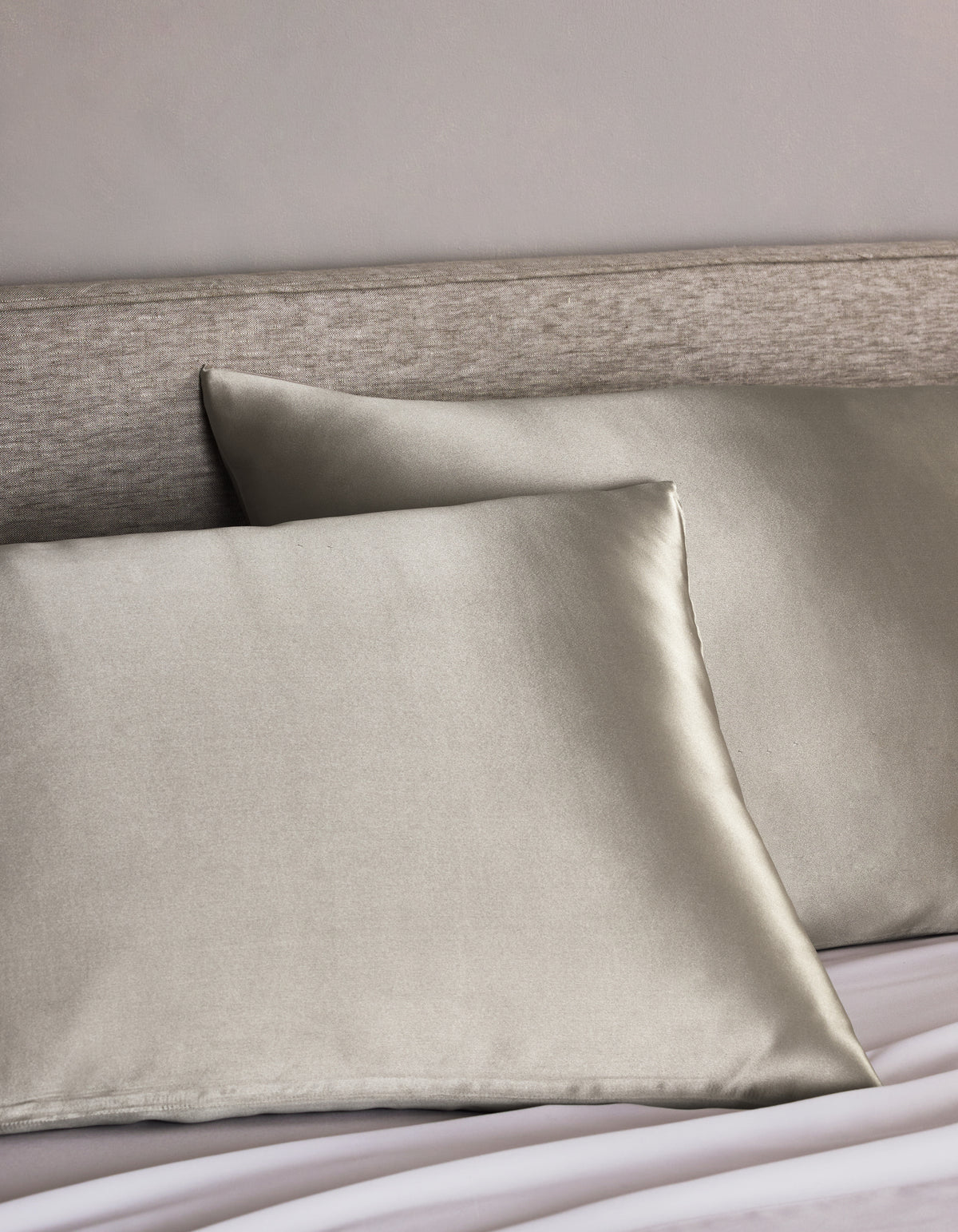 Two Cozy Earth Silk Pillowcases in light gray are arranged on a bed with a light beige upholstered headboard, adding a smooth, shiny, and luxurious touch to the bedding.