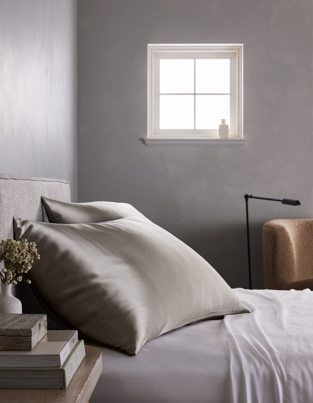 A neatly made bed features large, smooth Cozy Earth Silk Pillowcases in silver-gray and crisp white sheets beside a small table with books and flowers. A soft chair, lamp, and window on a gray wall complete the minimalist bedroom.